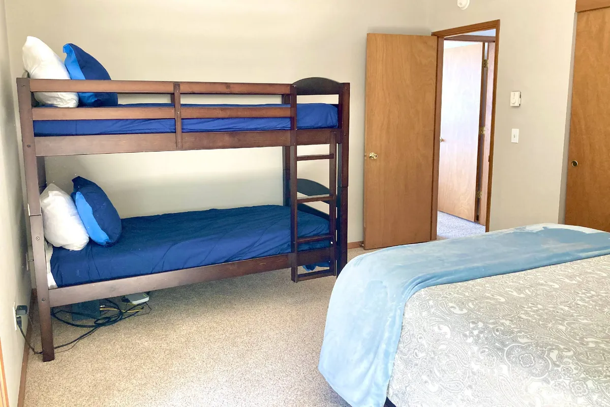 A welcoming guest bedroom with a queen bed, cozy bunk beds, and an open door leading toward the deck. The space is inviting and family-friendly, ideal for children or friends, and filled with natural light from the deck access.