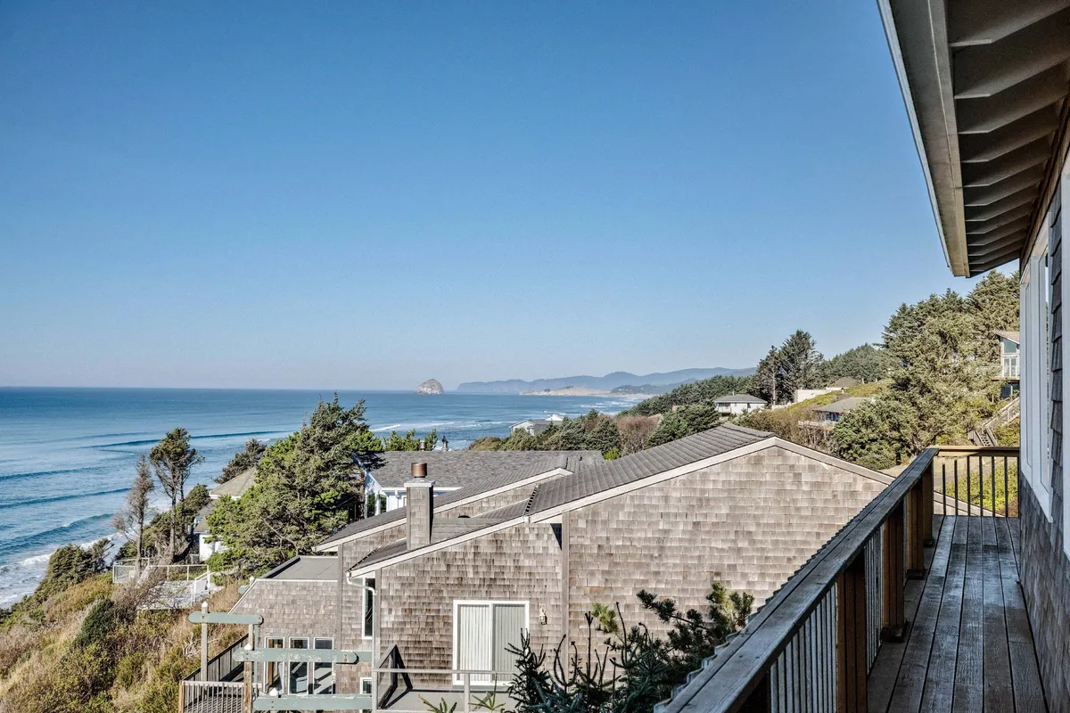 A wooden deck overlooking the ocean, with comfortable patio furniture strategically placed to enjoy the stunning sunset views. The scene includes tranquil ocean waves and distant cliffs, evoking the perfect setting for relaxing, wildlife watching, or stargazing after dark.