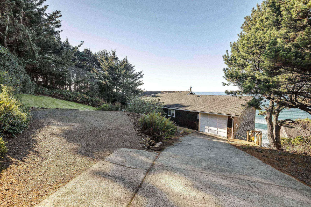 An exterior shot featuring the home's driveway leading to the house, flanked by trees and offering glimpses of the ocean in the distance. The peaceful, welcoming mood suggests a retreat where guests reconnect with both each other and the natural seaside environment.