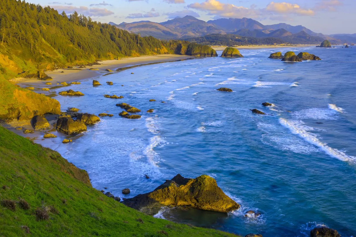 A panoramic scene featuring a rugged, rocky coastline, gentle ocean waves, and distant mountains under a calm sky. The image evokes a sense of peaceful adventure and is ideal for hiking, beachcombing, photography, and spotting wildlife.