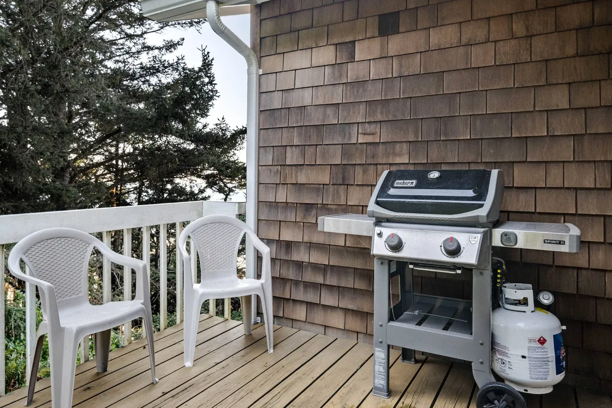 A spacious wooden deck with comfortable seating and a grill, framed by gentle coastal light. The view from the deck suggests it's a perfect spot for outdoor dining and socializing, with hints of ocean in the atmosphere beyond.