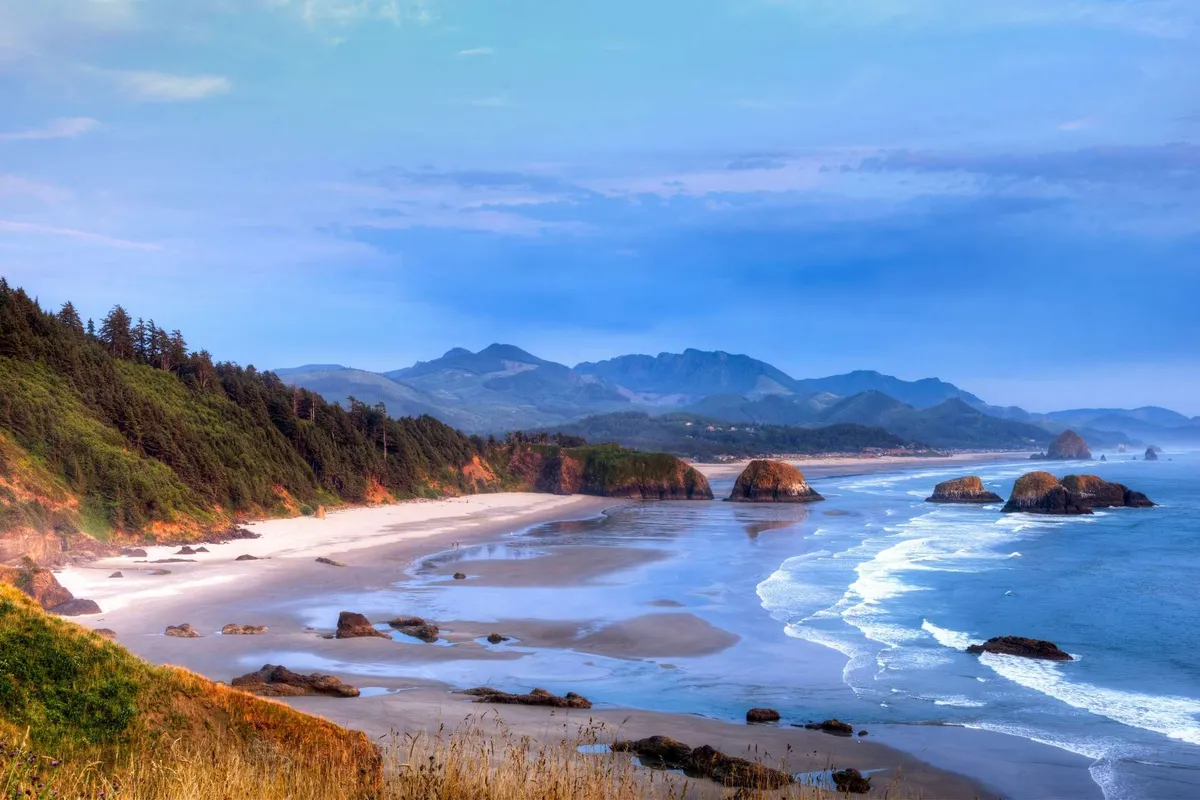 A wide view of the sandy beach, distant mountains, dramatic rock formations, and gentle ocean waves. The scene is tranquil, perfect for beachcombing, picnicking, or photographing sunsets and natural beauty.
