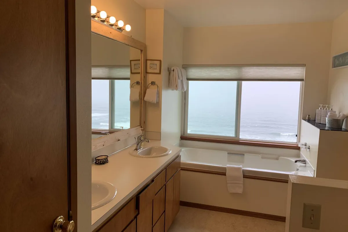 A spacious bathroom featuring a large window with ocean views, a double-sink vanity, and a luxurious bathtub. The setting is bright and tranquil, emphasizing a spa-like atmosphere perfect for unwinding while soaking and gazing out at the Pacific.