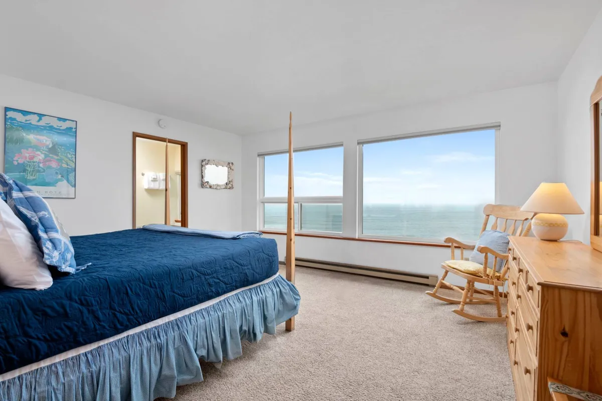 A serene master bedroom with blue bedding, large windows that overlook the ocean, a comfortable rocking chair, and a dresser. The room is bathed in natural light, offering direct views of the sea and creating a tranquil retreat.