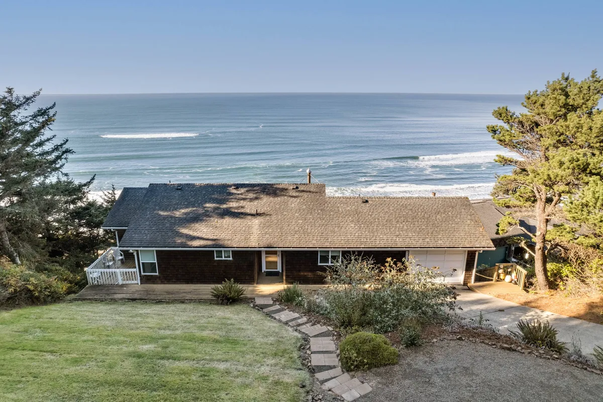 The house's facade and landscaped yard open to sweeping ocean views, with a welcoming deck and pathway inviting relaxation. The setting glows under the warm light of late afternoon, hinting at memorable sunsets and peaceful evenings.