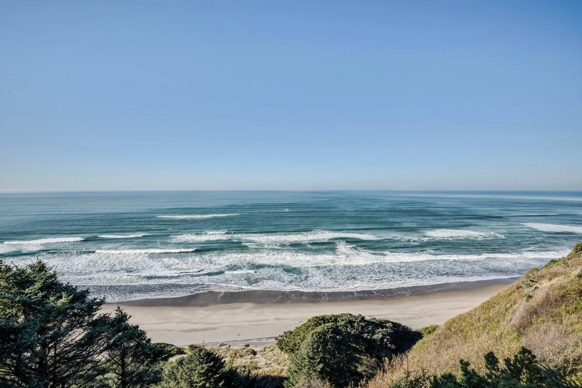 An expansive sandy beach meets rolling ocean waves under a tranquil sky, with dramatic cliffs and lush green foliage framing the coastline. There are footprints in the sand and plenty of open space, hinting at families building sandcastles, walking, or enjoying a picnic by the seaside.