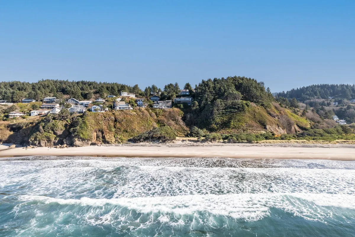 A sweeping view of a serene beach bordered by green hills, scattered houses on a nearby cliff, and gentle ocean waves rolling ashore. The image captures the natural beauty and tranquility of the coastal environment, perfect for activities like hiking, beachcombing, and wildlife watching.