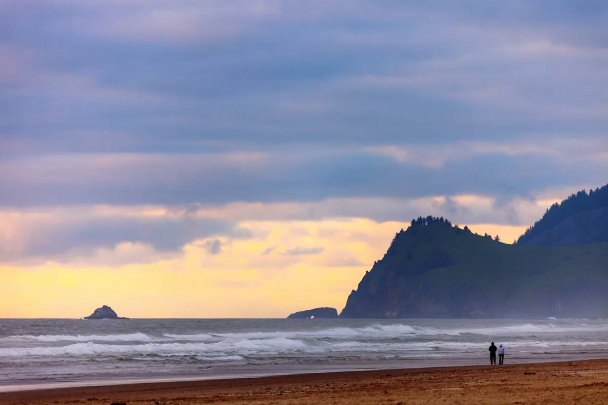 A couple walks hand in hand along a peaceful sandy beach with gentle ocean waves rolling in, distant cliffs framing the horizon, and the sky tinged with sunset colors. The tranquil setting evokes romance and connection with nature.