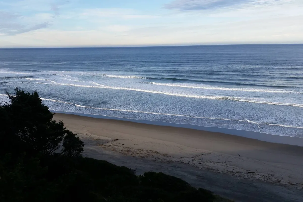 A calming beach scene featuring gentle ocean waves, a sandy shoreline, and a distant horizon. People are seen walking along the beach and watching surfers, evoking an active yet peaceful seaside atmosphere. The rising sun casts a warm light over the water.