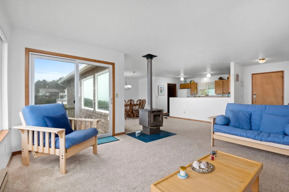 This inviting interior scene showcases a living/dining area with large windows revealing stunning ocean views. The space includes a dining table for shared meals, blue sofas for lounging, and a decorative wood stove, all set in a homely, welcoming environment.