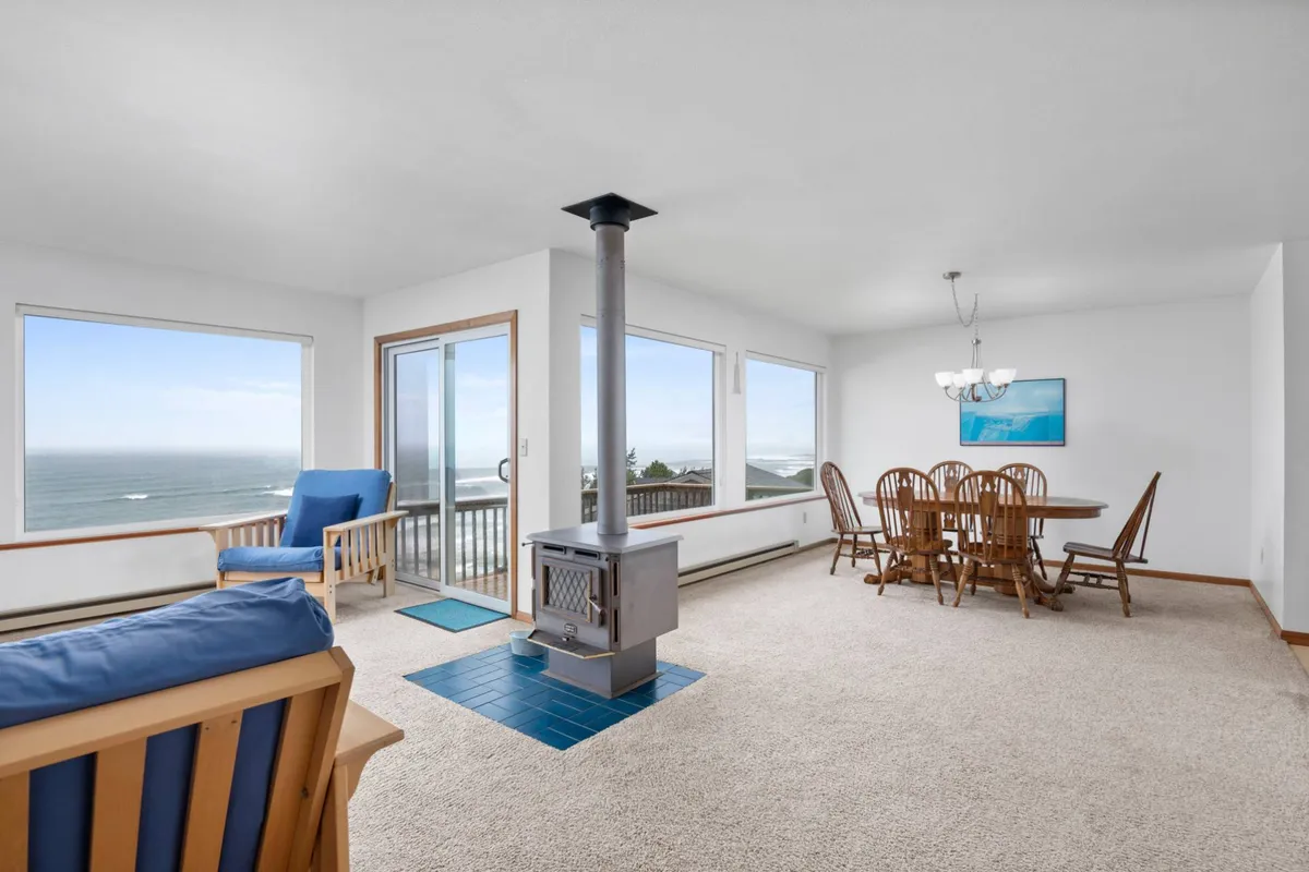 This image features a spacious interior with large windows framing sweeping ocean views. Central to the room is a sizable wooden dining table, set close to the windows for a scenic dining experience. The room includes a wood stove for rustic charm, and the overall décor is cozy and inviting, emphasizing family gatherings and relaxation.