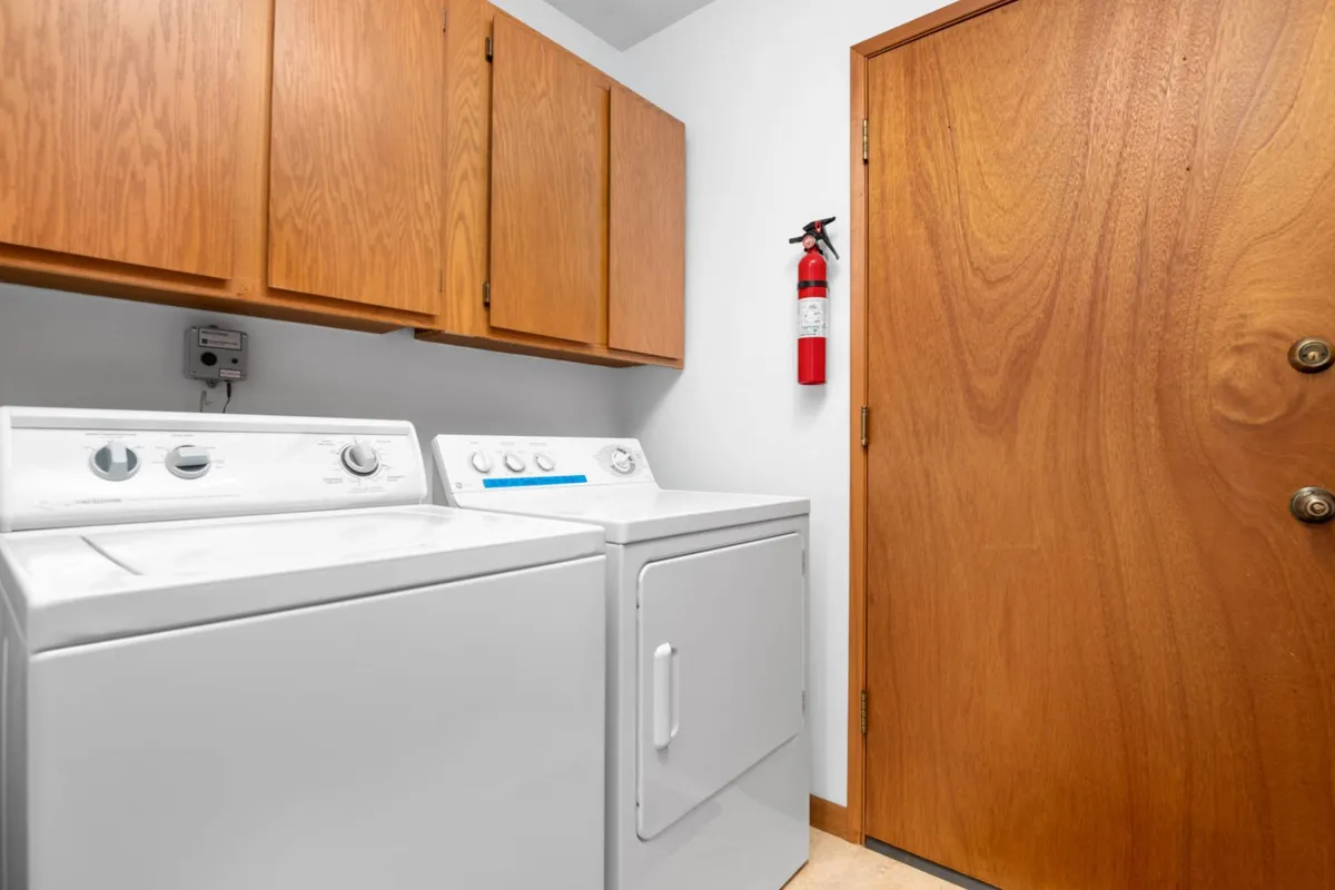 A bright and tidy laundry area featuring a washing machine and dryer set underneath wooden cabinets, with a front door and fire extinguisher nearby. The setup is functional and clearly dedicated to laundry tasks, emphasizing convenience and organization.