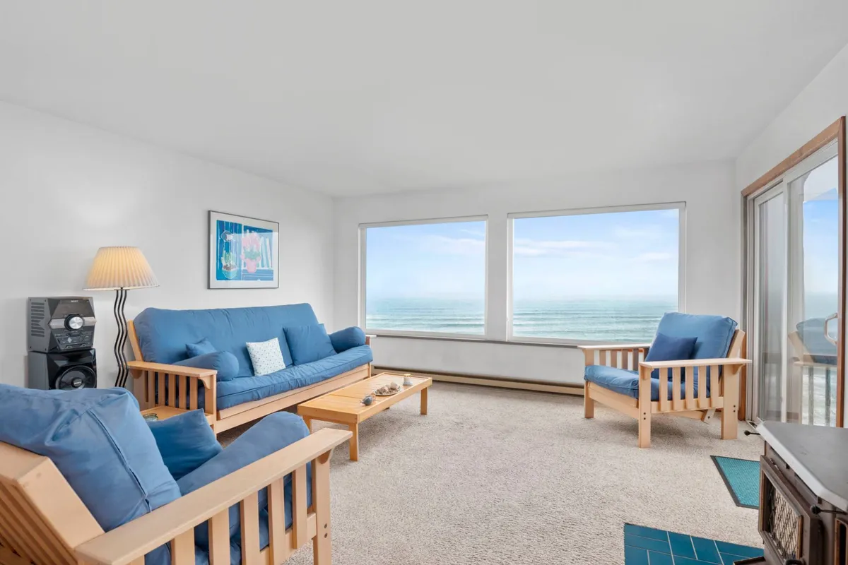 A welcoming living area is shown, with comfortable blue sofas arranged to face out toward expansive picture windows that look onto the ocean. A wooden coffee table sits at the center, while natural light fills the space, creating a tranquil, inviting atmosphere ideal for relaxation and conversation.